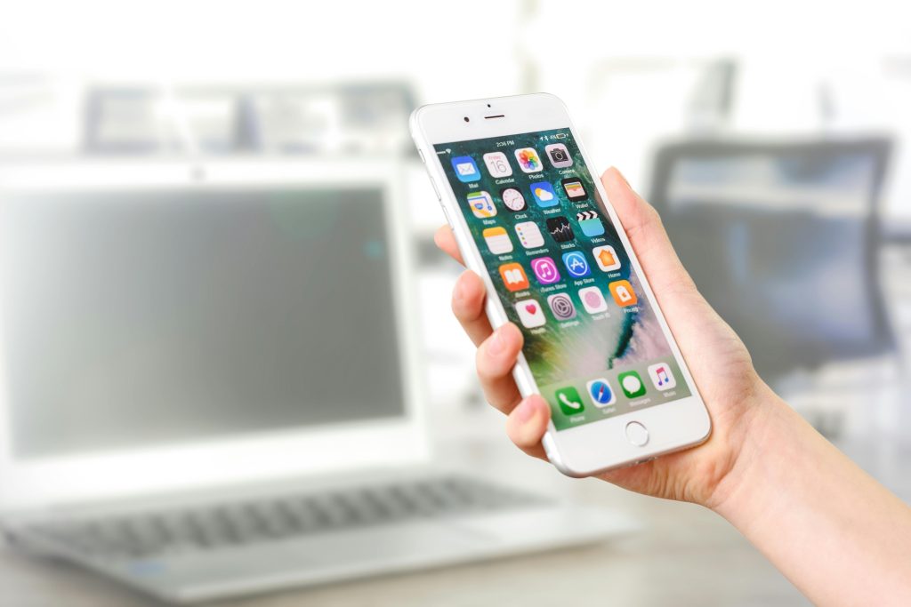 pexels-photo-887751-887751 Close-up view of a smartphone displaying apps, held by a hand, with a blurred laptop in the background.